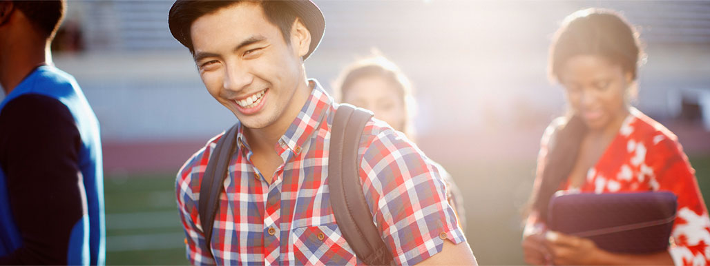 Asian male student wearing a blue, red, and white plaid shirt and a hat carrying a backpack smiling