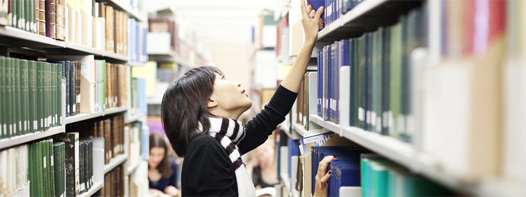 Asian student looking through books on shelf in library