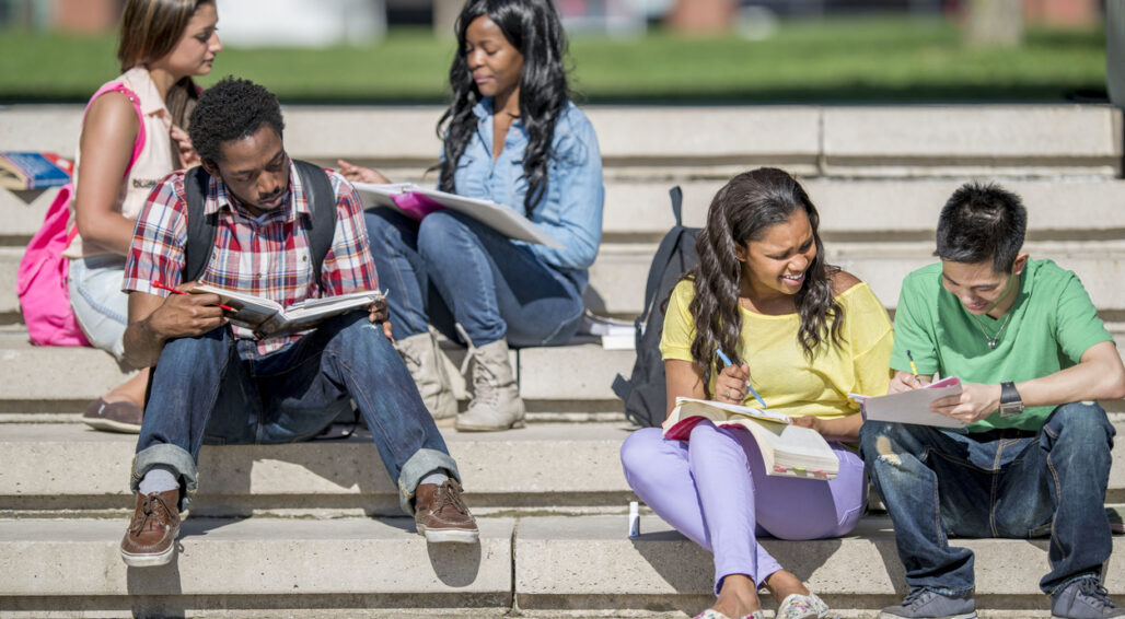 A multi-ethnic group of college age students are studying together outside on cement steps on a beautiful sunny day.
