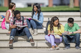 A multi-ethnic group of college age students are studying together outside on cement steps on a beautiful sunny day.