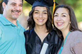 Graduate in black cap and gown holding diploma with mom and dad