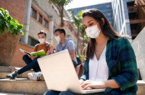 Female young student working on laptop sitting around the college campus and everyone around wearing protective face masks