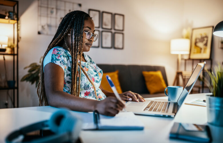 Black woman writing at a desk in front of a laptop